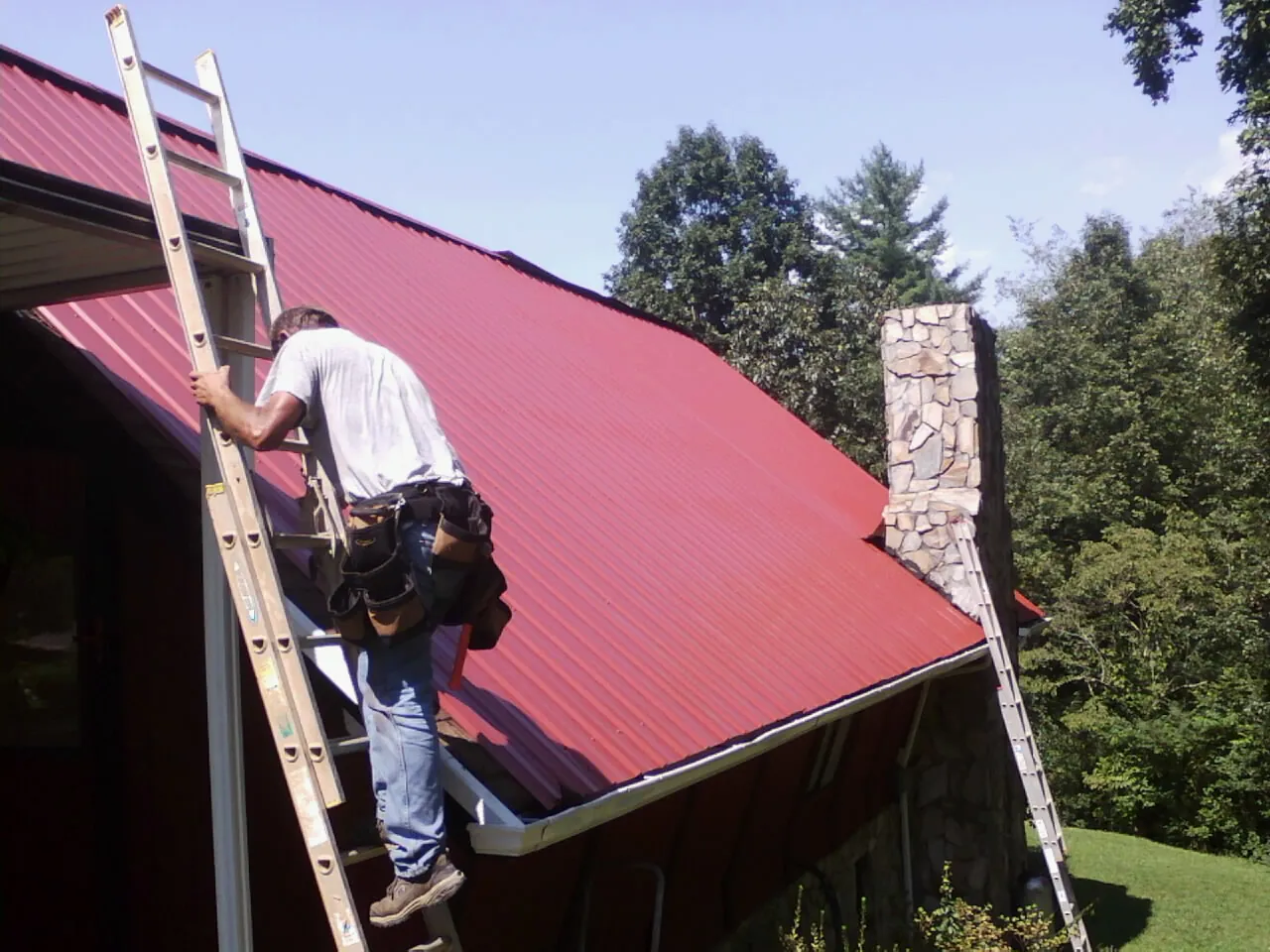 Roofer on a ladder beside a red metal roof and stone chimney — Soffit & Fascia Repair by Roofing Brothers in O Fallon